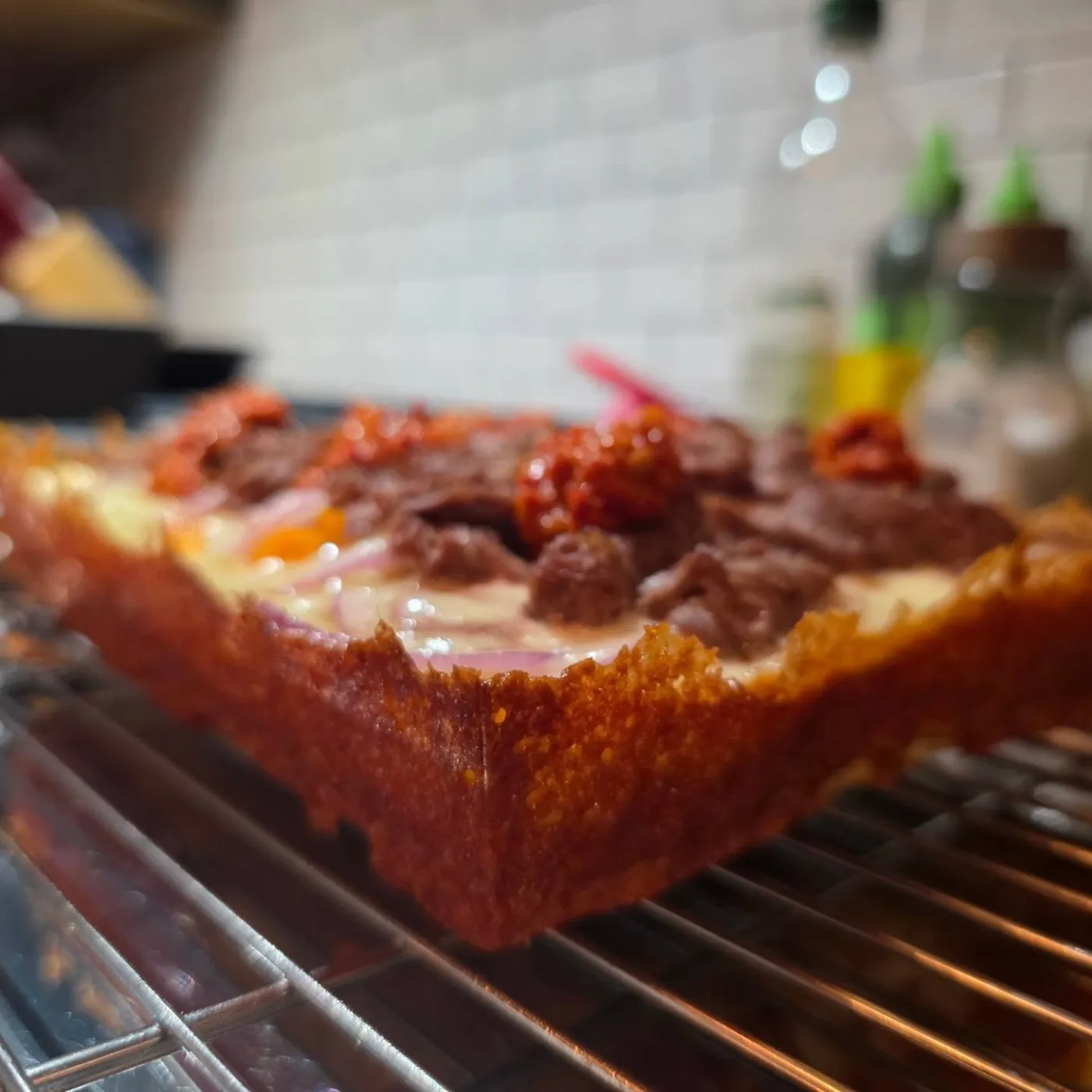 Close-up of a Detroit-style pizza's deep golden, caramelized frico crust edge, topped with beef and red onion, on a cooling rack